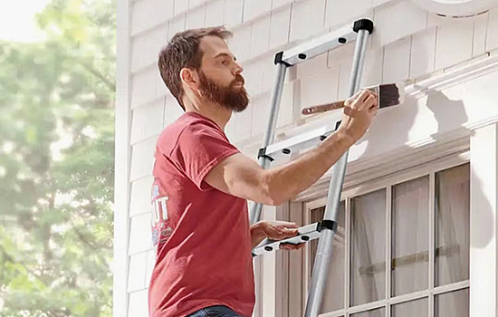 A man is standing on a telescopic extension ladder and painting the wall